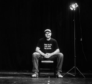 Black and white photograph of Alastair Moock, Williams class of 1995, seated on a chair on stage