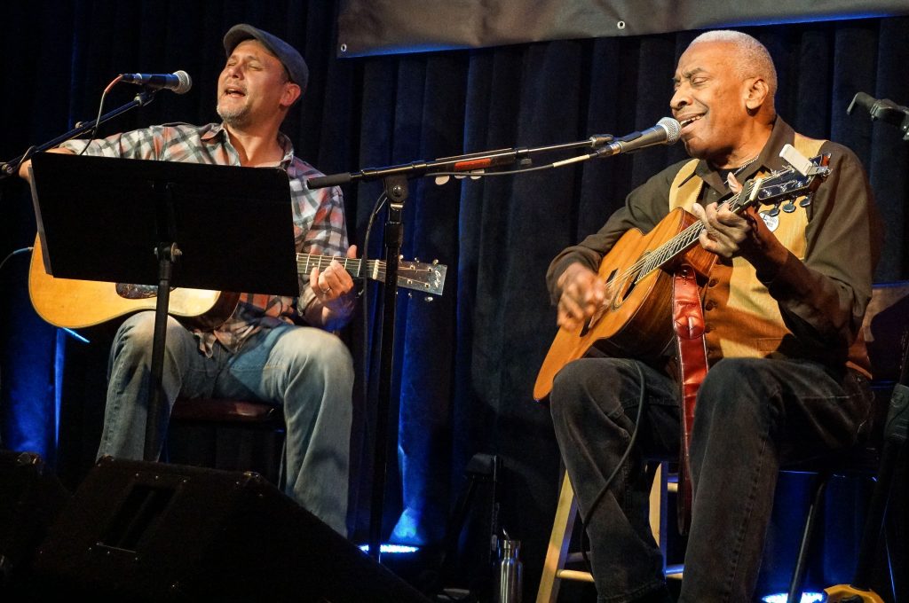 Alastair and Reggie 2a Two musicians, Alastair Moock, Williams class of 1995, and his friend and collaborator Reggie Harris play guitar, seated, on stage.