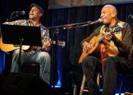 Two musicians, Alastair Moock, Williams class of 1995, and his friend and collaborator Reggie Harris play guitar, seated, on stage.