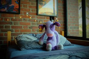 A knit stuffed purple cow sitting on a bed.
