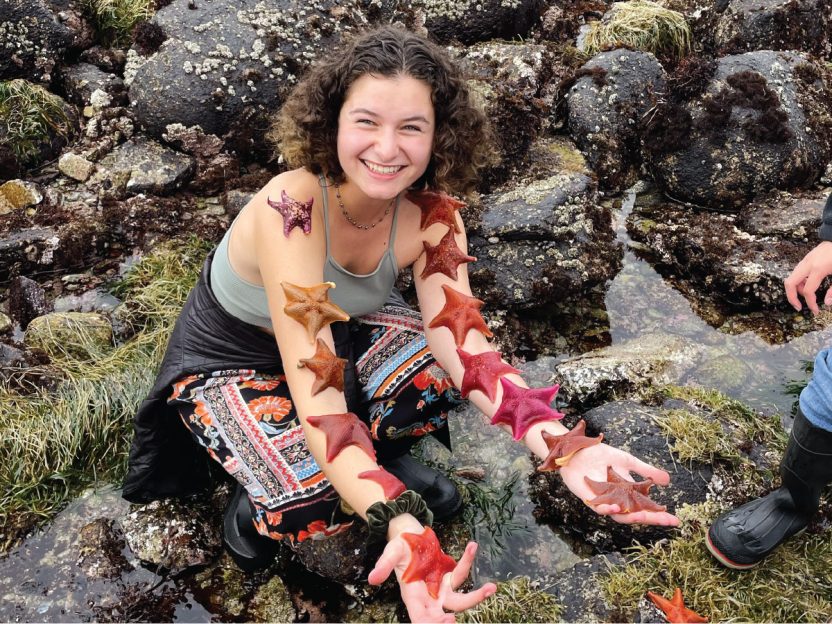 Photo of a student kneeling by the water with starfish along each of her arms.