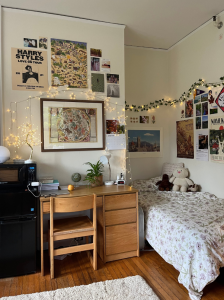 A corner of a college dorm room shows a bed and a desk and is decorated with posters, lights, and vines.