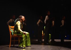 A performer in a shiny lime green costume sits onstage in a chair, leaning forward slightly and shouting while her squad, clad in black and white, stands beside her with arms folded behind their backs.