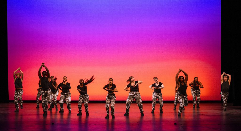 Team3_PS_DSC_0334-Enhanced-NR-1 A group of dancers perform a step routine against a magenta and orange background. They are in a crouching position, some with arms raised over their heads and some with arms folded in at chest height.