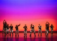 A group of dancers perform a step routine against a magenta and orange background. They are in a crouching position, some with arms raised over their heads and some with arms folded in at chest height.