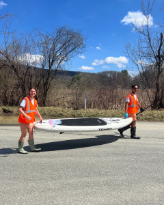 Two students in orange vests carry a white kayak down a road with trees in the background