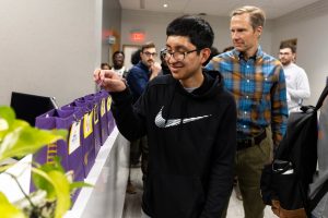 Two people place slips of paper into a row of purple paper bags, each with a business name and logo attached.