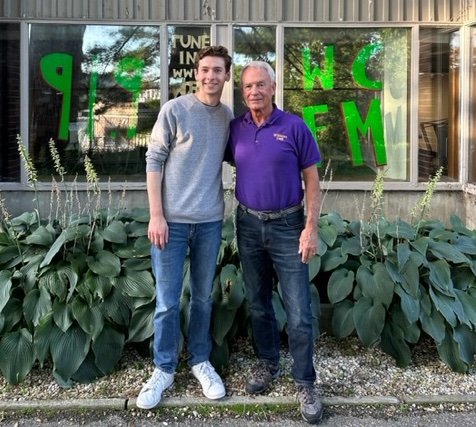 Two people stand outside a building with gray siding and large windows. Large green letters in a window behind them say 91.9 WCFM, and smaller white letters say Tune in with a URL (not entirely visible in the photo).